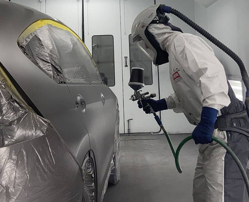 Mechanic in white hazmat suit spray painting a sedan with silver paint in an auto body garage